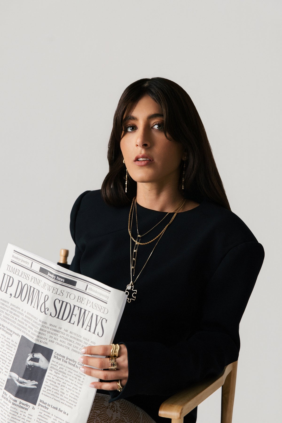 A woman with dark hair (Jess Klein, founder) sits in a wooden chair against a light gray background, wearing a black long-sleeve top and layered gold necklaces. She is holding a newspaper.